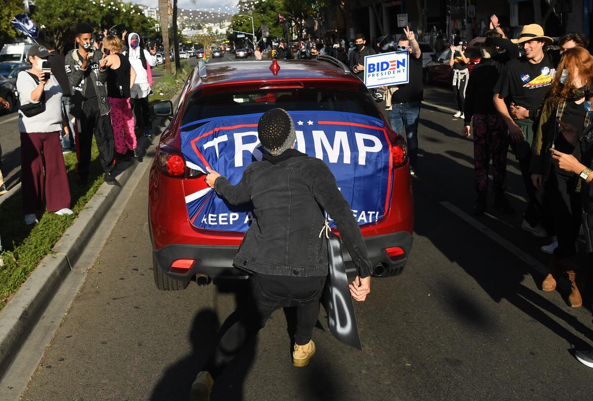 A man rips a blue Trump 2020 flag off the back of a red car driving between crowds of Biden supporters