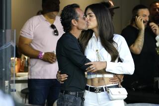 A middle-aged man holds and kisses a younger woman in a luxury box before a sporting event