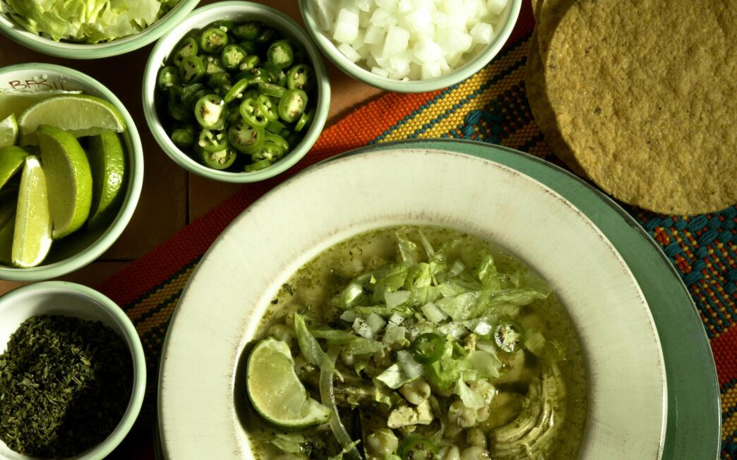 Pozole verde (green pozole) with condiments bottom left clockwise: oregano, lime, lettuce, serrano chile, onion and tostadas.