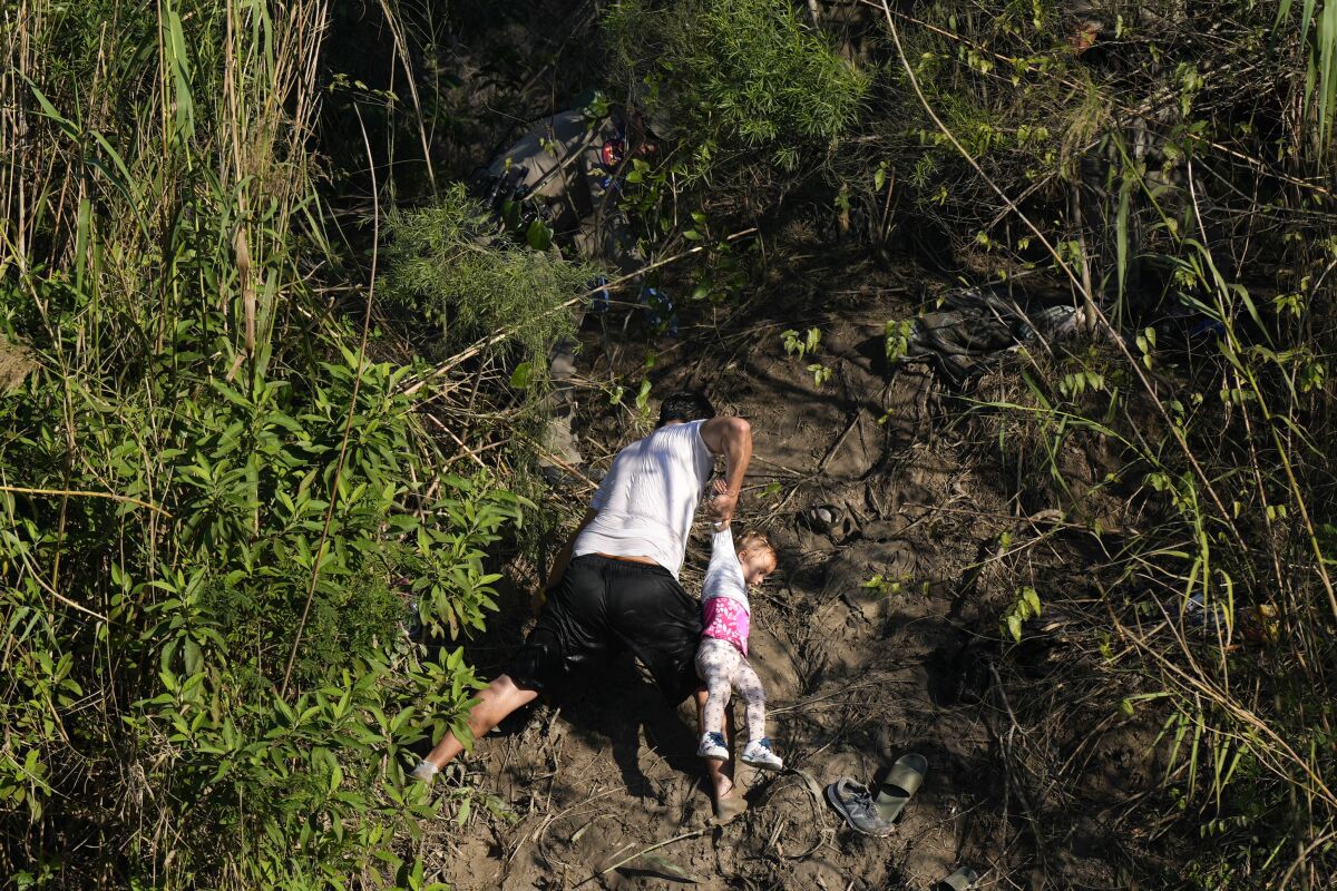 Bir adam, çamurlu bir nehir kıyısına tırmanırken bir çocuğu kolundan tutuyor. 