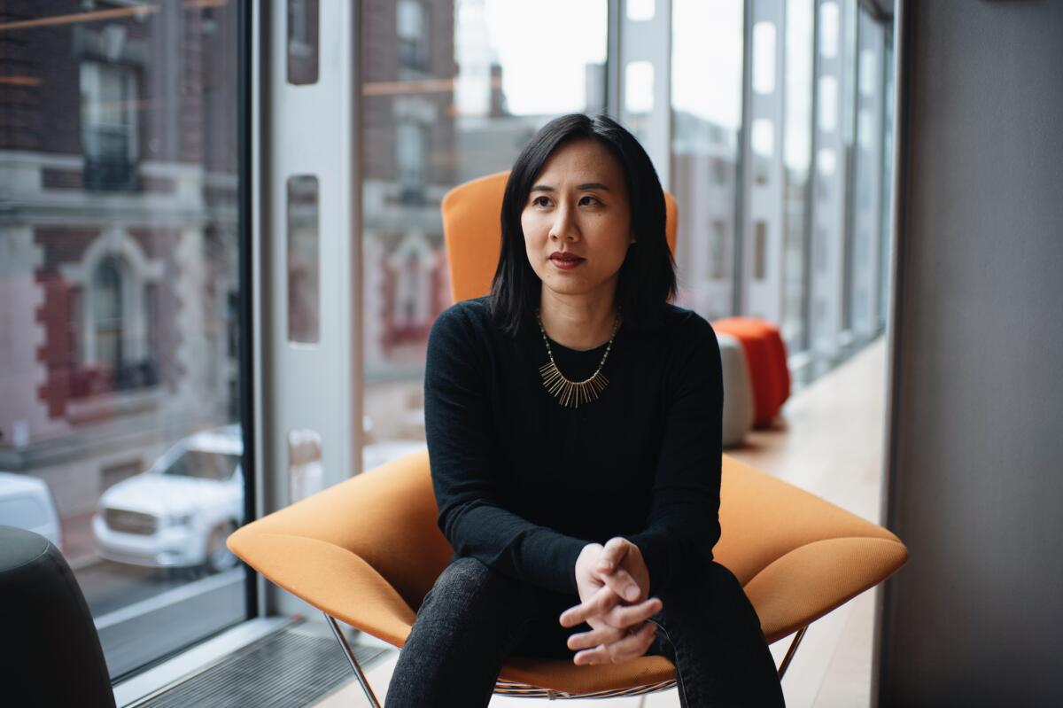 A woman sits in a modern, angular chair in front of glass walls with city buildings in the background.