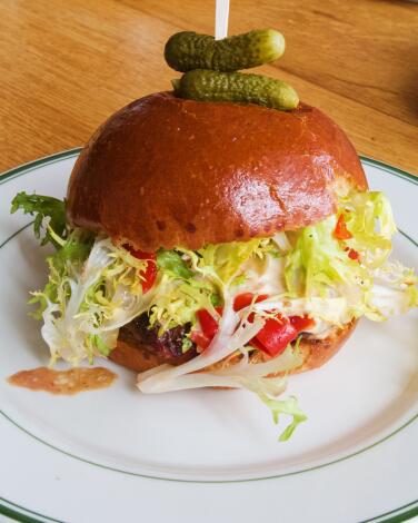 A vertical shot of Coucou's Burger Americaine, frisée peeking out from beneath a brioche bun that's skewered with cornichons