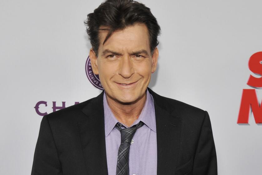 Charlie Sheen smiles and poses while wearing a dark suit and light purple shirt with a gray patterned tie