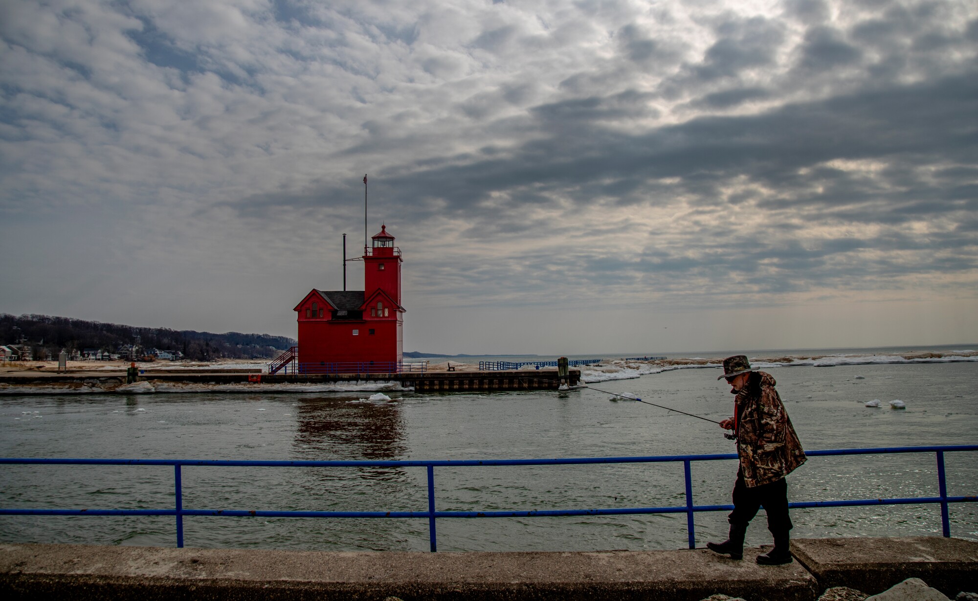A man with a fishing pole walks along the waterfront near a red lighthouse