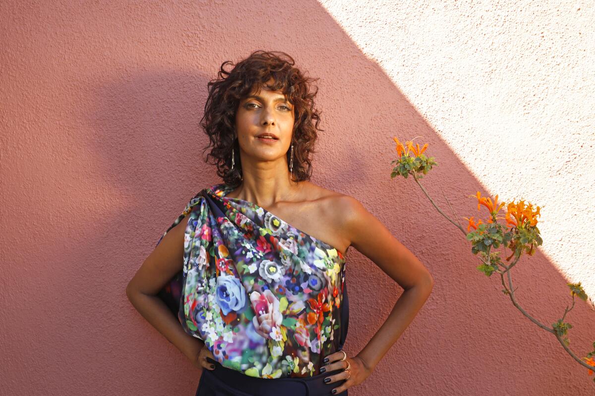  Poorna Jagannathan standing before a rose-colored wall in a floral one-shoulder blouse next to orange flowers.