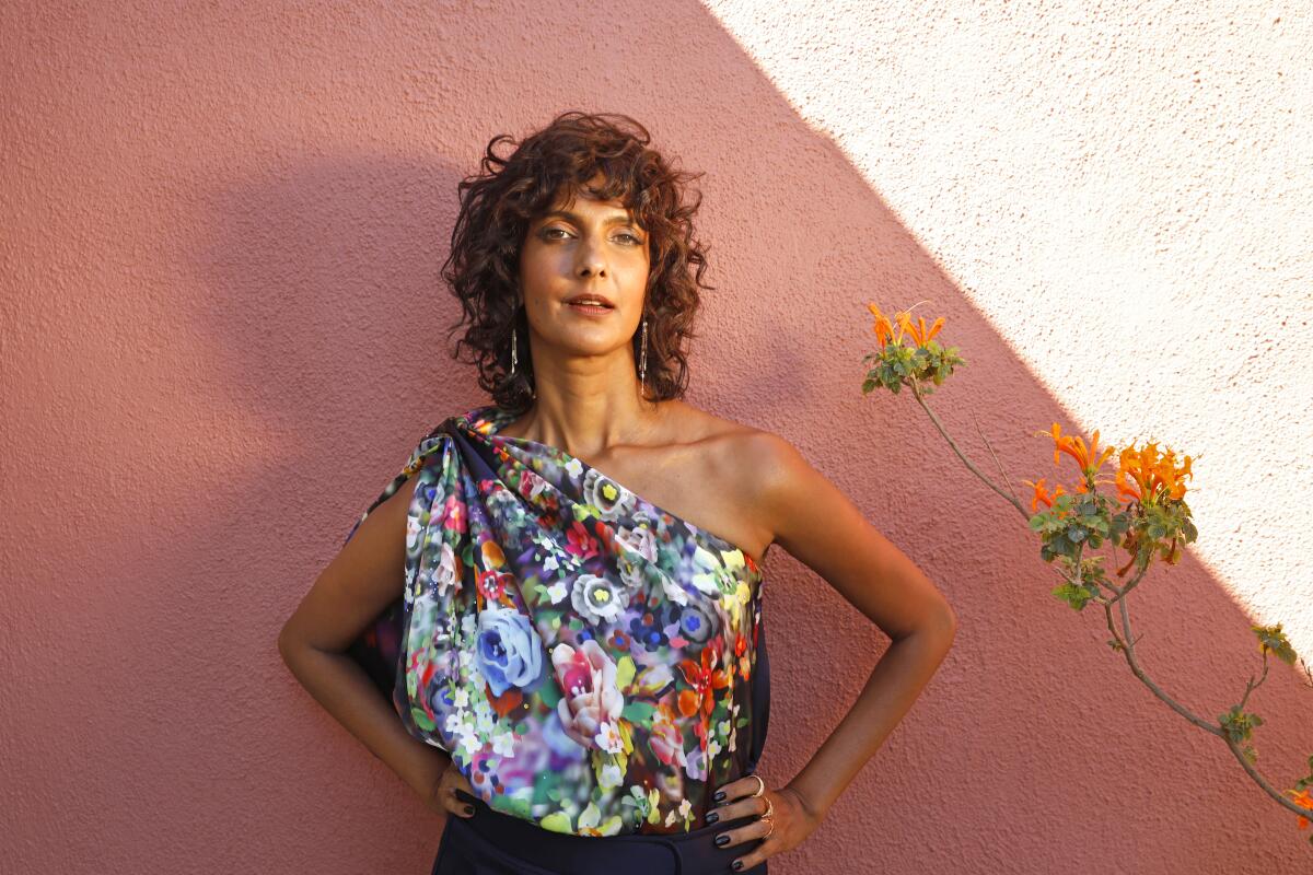Poorna Jagannathan standing before a rose-colored wall in a floral one-shoulder blouse next to orange flowers.