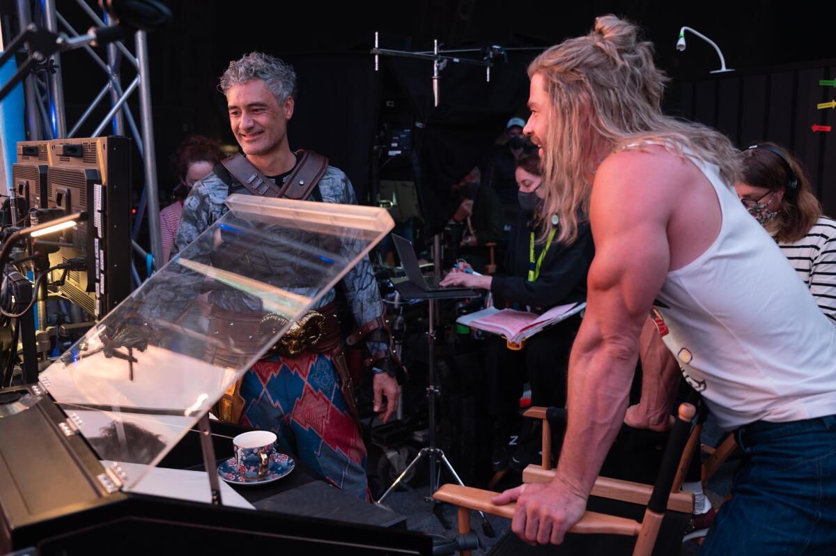 A man in a superhero costume and a long-haired man with muscly arms in a tank top talk on a movie set.