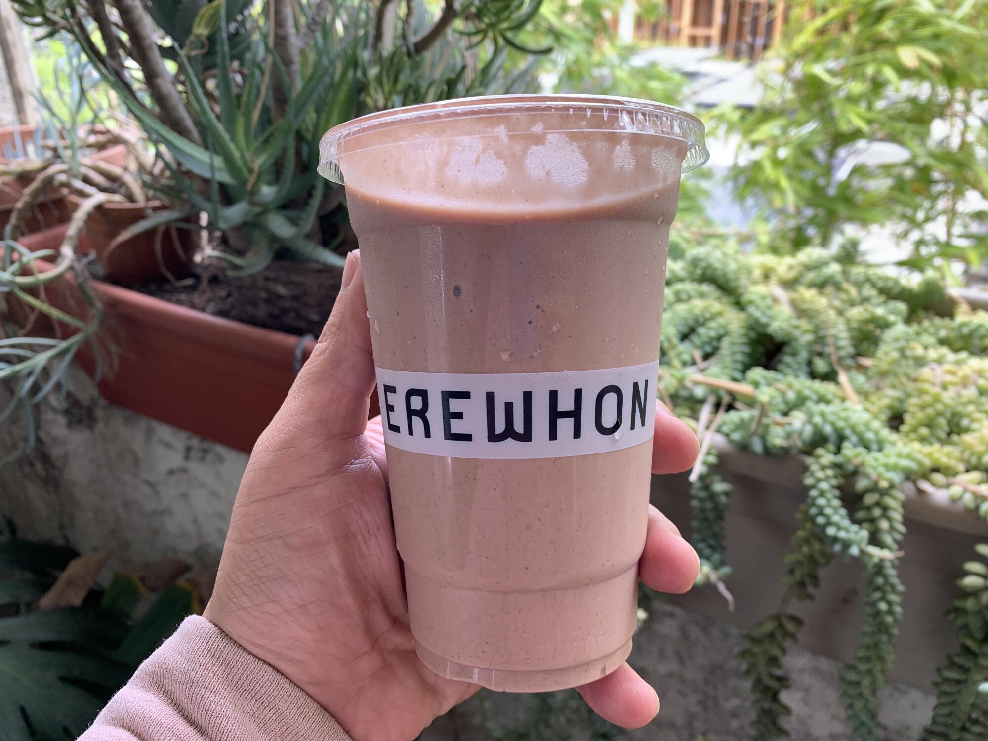 A hand holds a brown smoothie in a plastic cup that says Erewhon.