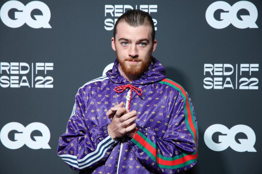 A man in a shaved head wearing a purple patterned jacket with Adidas and Gucci stripes on each sleeve posing