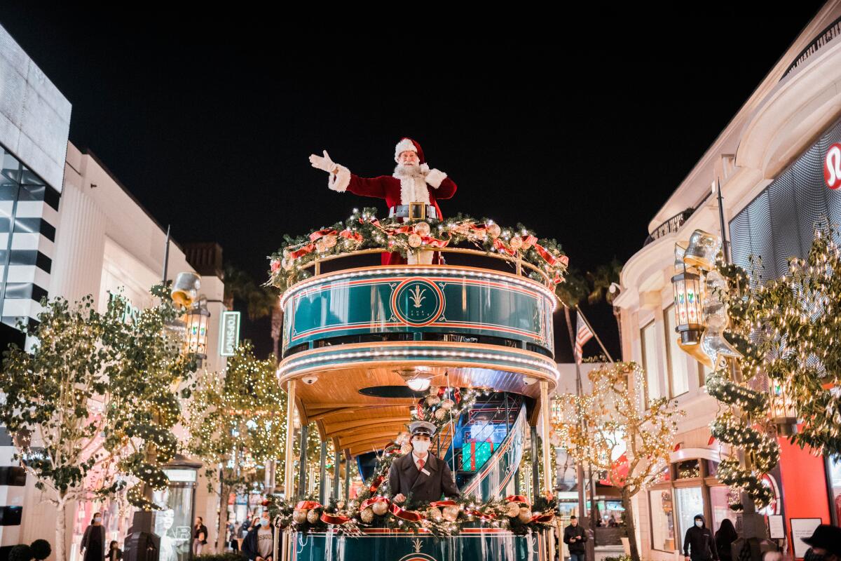 Santa stands atop a trolley decorated with holiday lights in the middle of the Grove shopping center