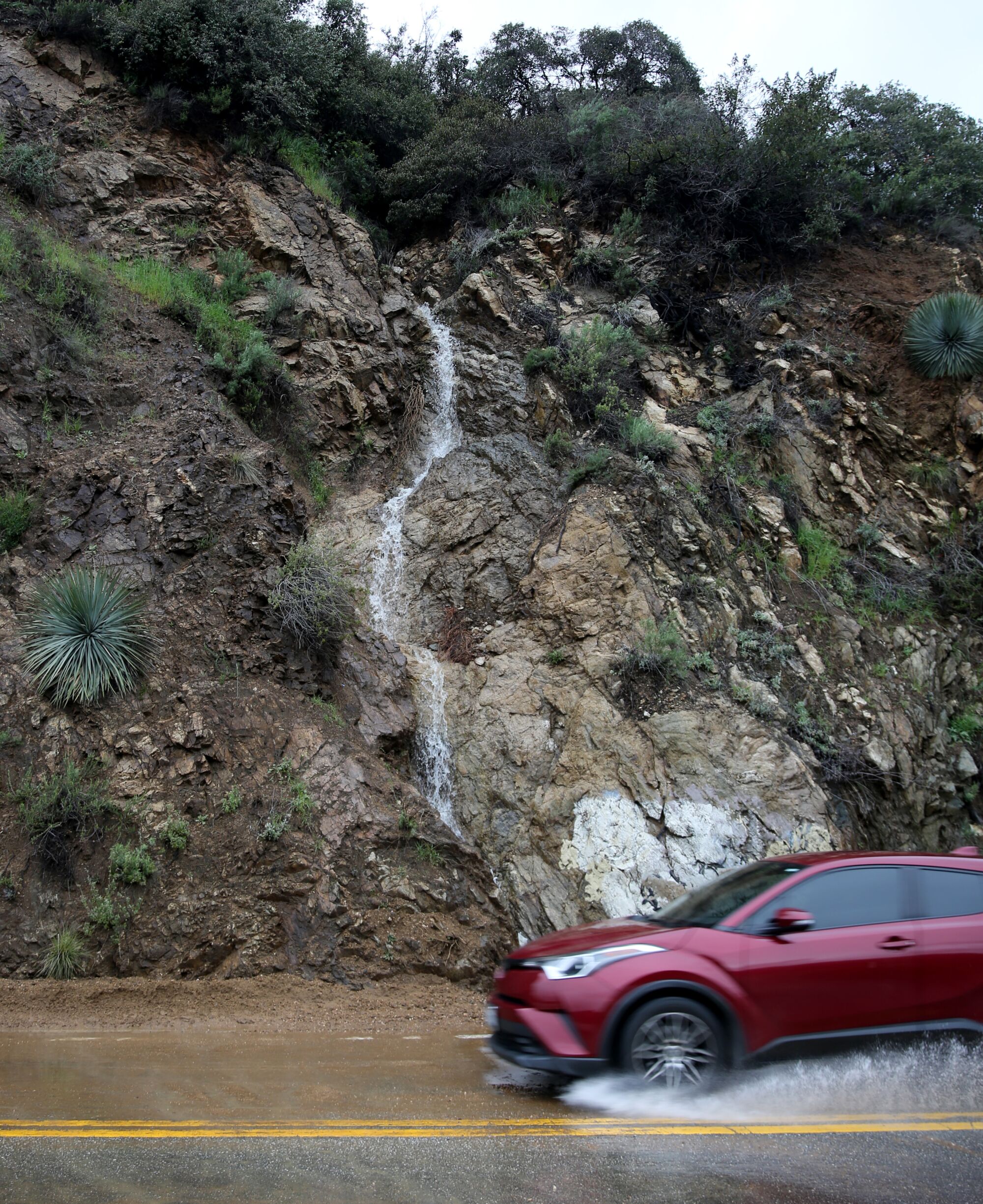 A watefall pours onto a roadway as a car splashes through a puddle.