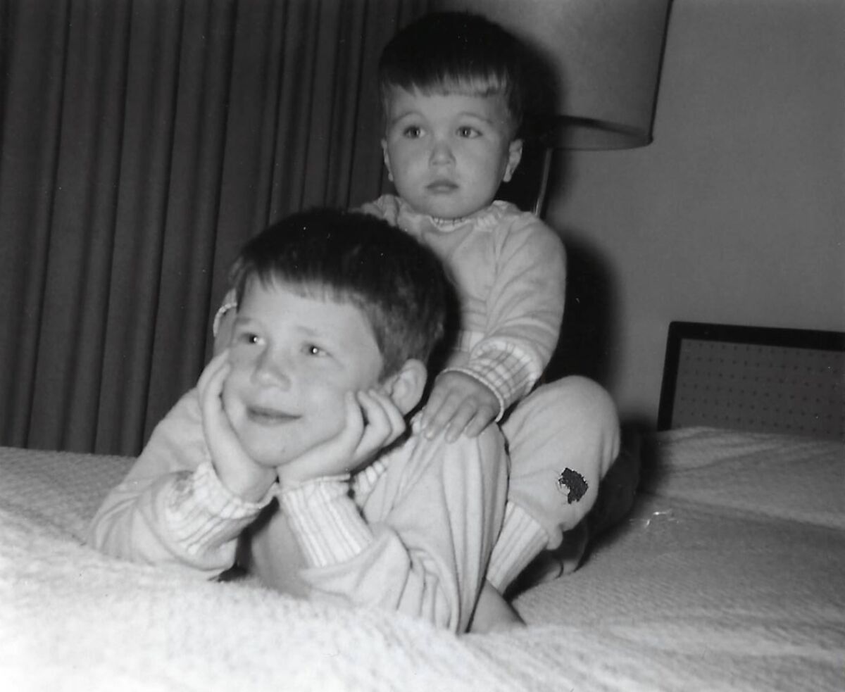 Two young boys, one lying on his stomach on a bed, the other sitting on his back. 