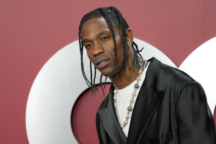 Travis Scott poses with braided her and in a black blazer and beaded necklace