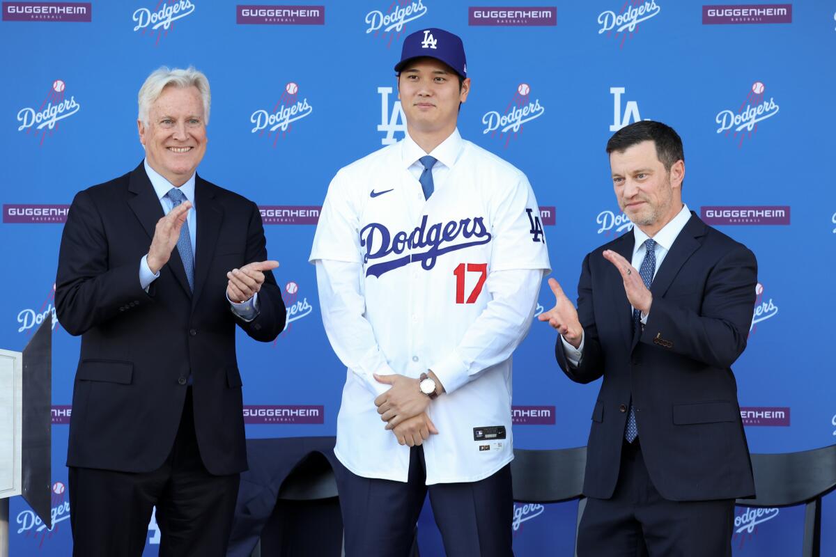 Shohei Ohtani stands between two men in suits and ties. He wears a Dodgers jersey and cap.