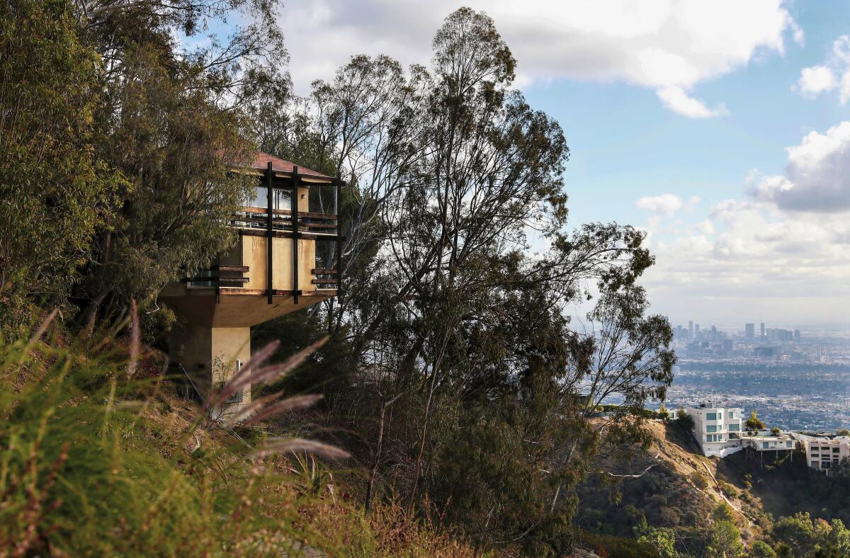 A house is perched on a hillside amid trees. In the background is a view of the Los Angeles skyline.