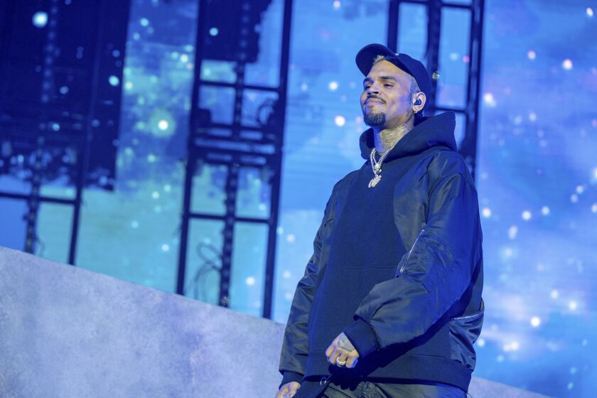 Man, Chris Brown, dressed on blue lit stage in black hat, jacket, hoodie and pants, smiling to audience 