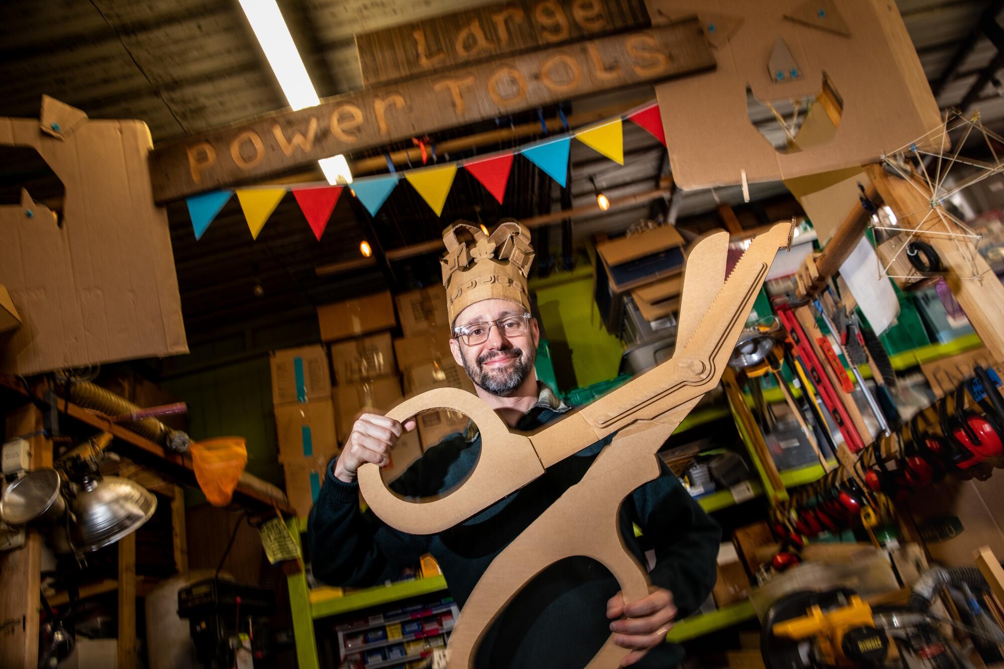 A man wearing a cardboard crown holds a large pair of scissors that is cut from cardboard.