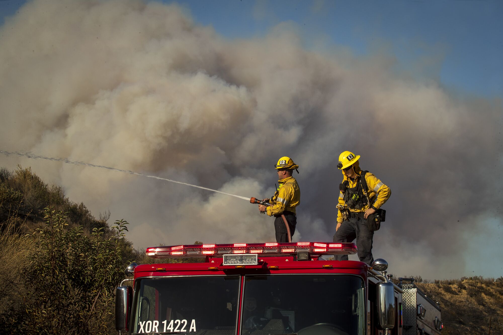 Firefighters on top of a fire engine, one of them using a hose to spray water on a hillside, with clouds of smoke behind them