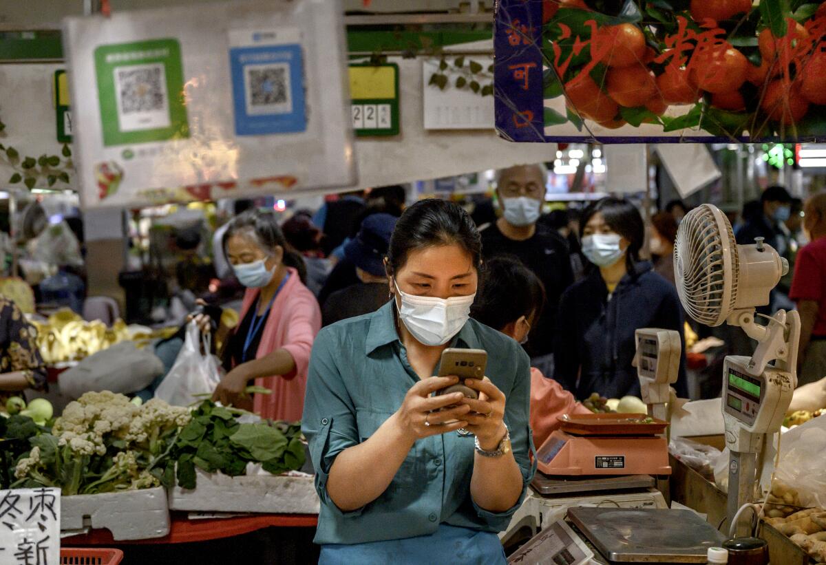 A masked woman looks at her cellphone as she stands under a QR code, amid produce and scales