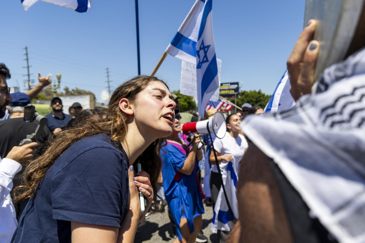 A young woman with long hair, wearing a blue T-shirt, stands near a blue-and-white flag and other people