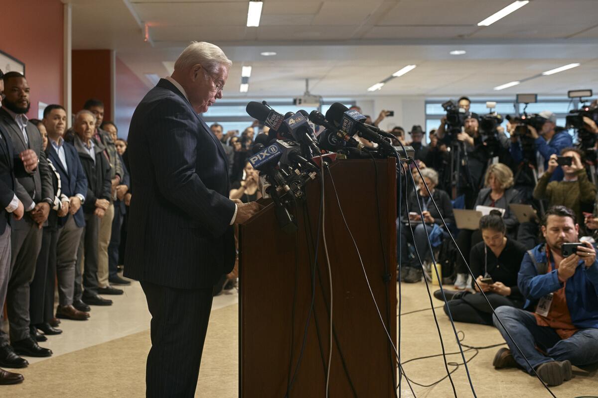 A man looking down, at a lectern, with reporters watching 