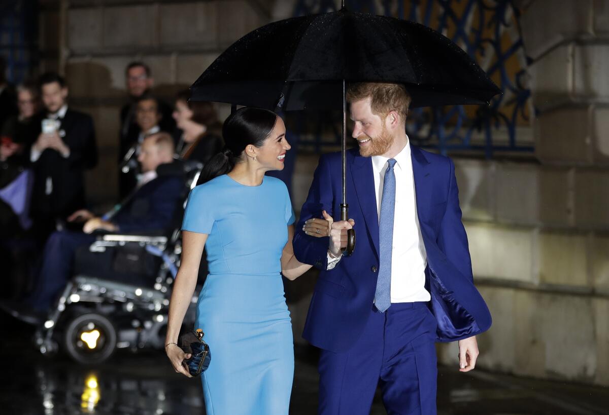 A dark-haired woman in a blue dress, left, and bearded man in blue suit and tie, smile as each other under an umbrella  