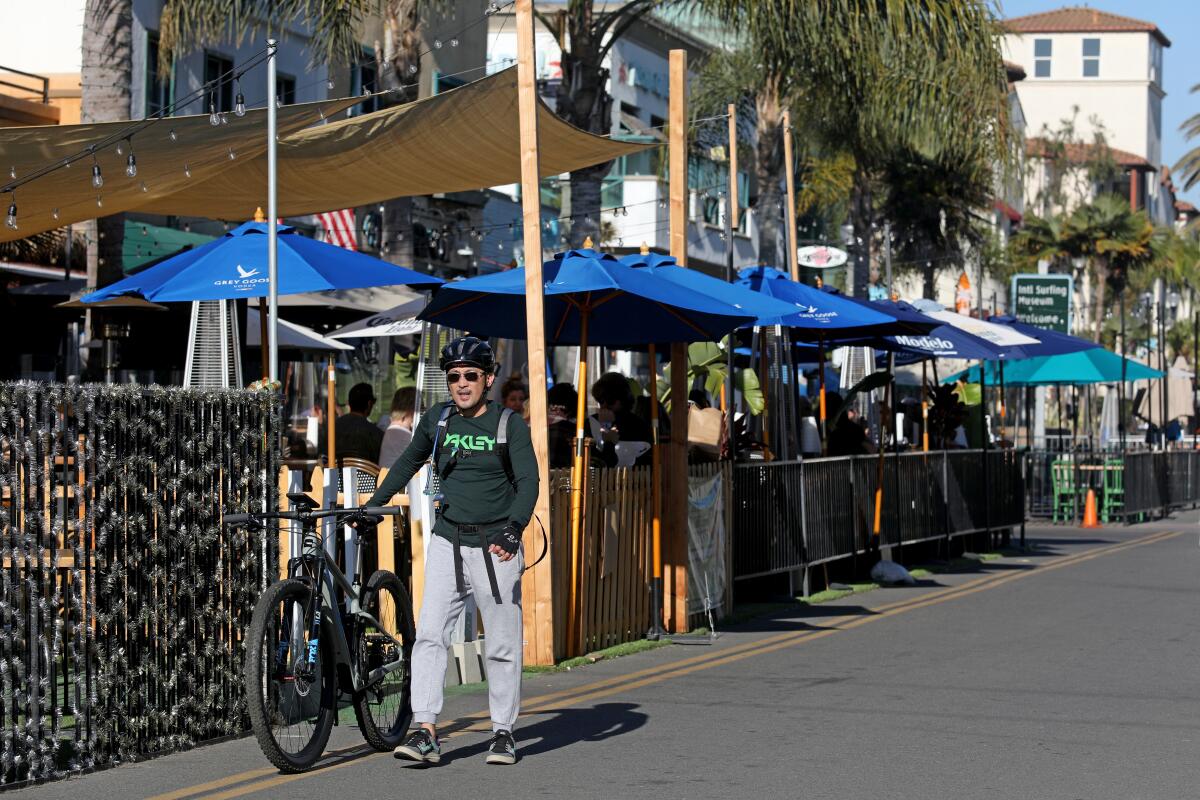 A man in a bike helmet walks near umbrellas covering sidewalk seating at a restaurant.