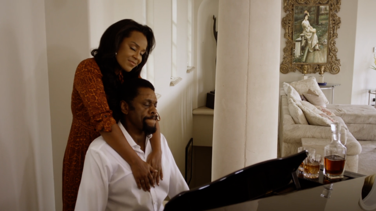 A standing woman drapes her arms around a man seated at a piano.