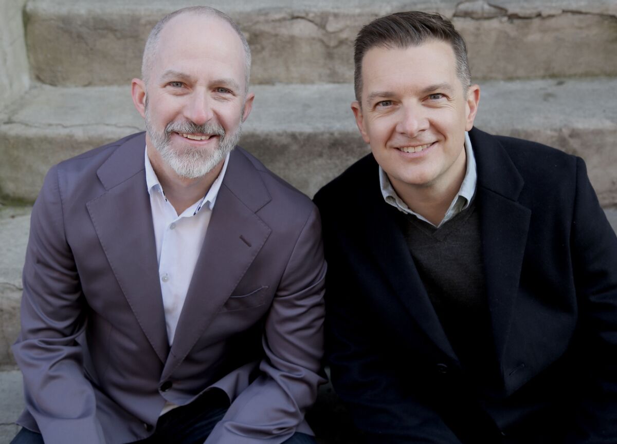 Two smiling men sitting on stone steps. One is white-bearded, in a grey jacket. The other: close-cropped, dark hair & jacket.