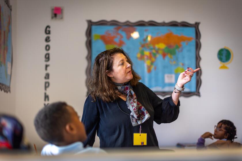 A teacher instructs students in a classroom with a geography display and a world map on the wall behind her.