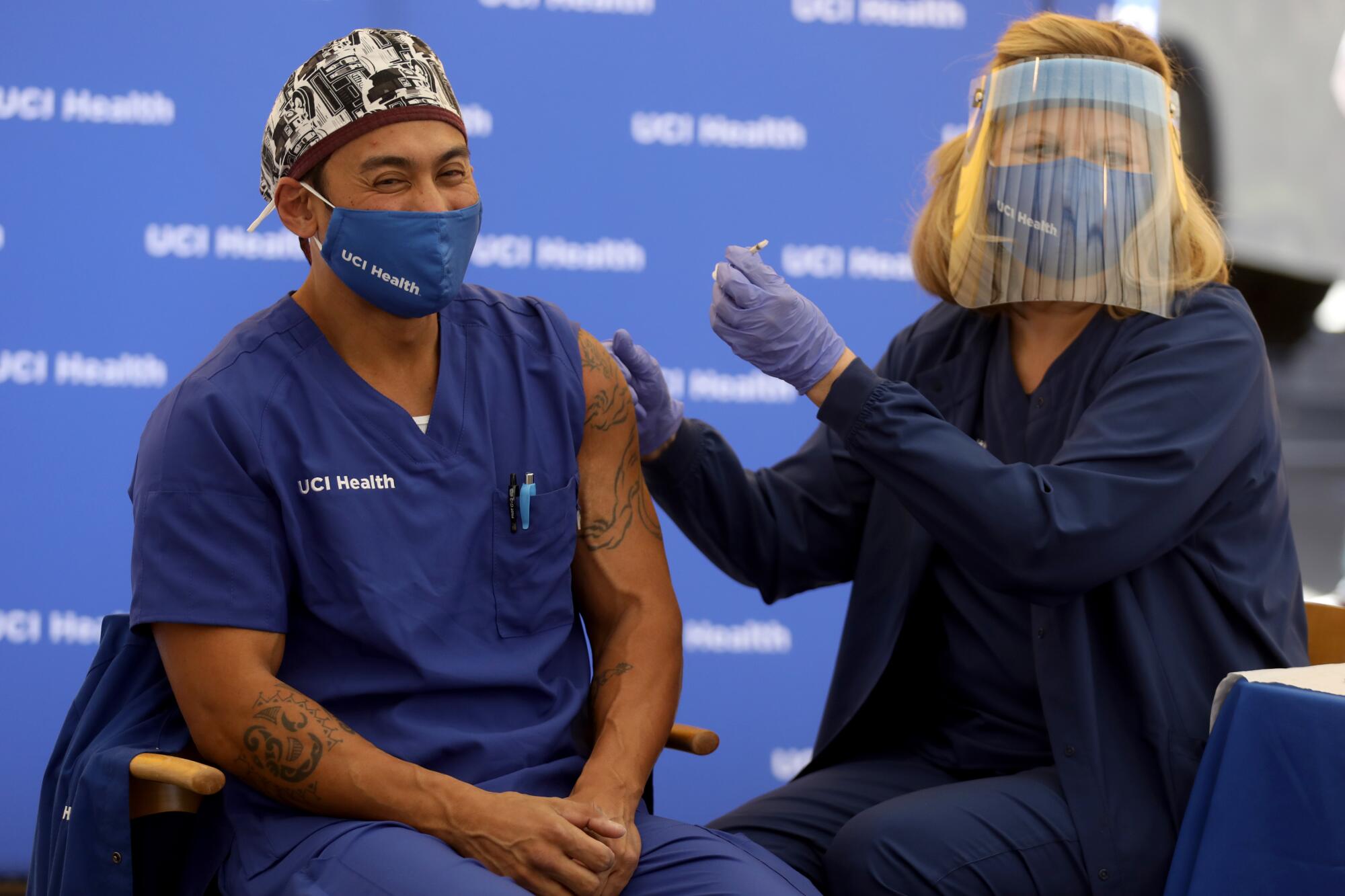 A healthcare worker dressed in scrubs receives a vaccine administered by a nurse wearing a face shield