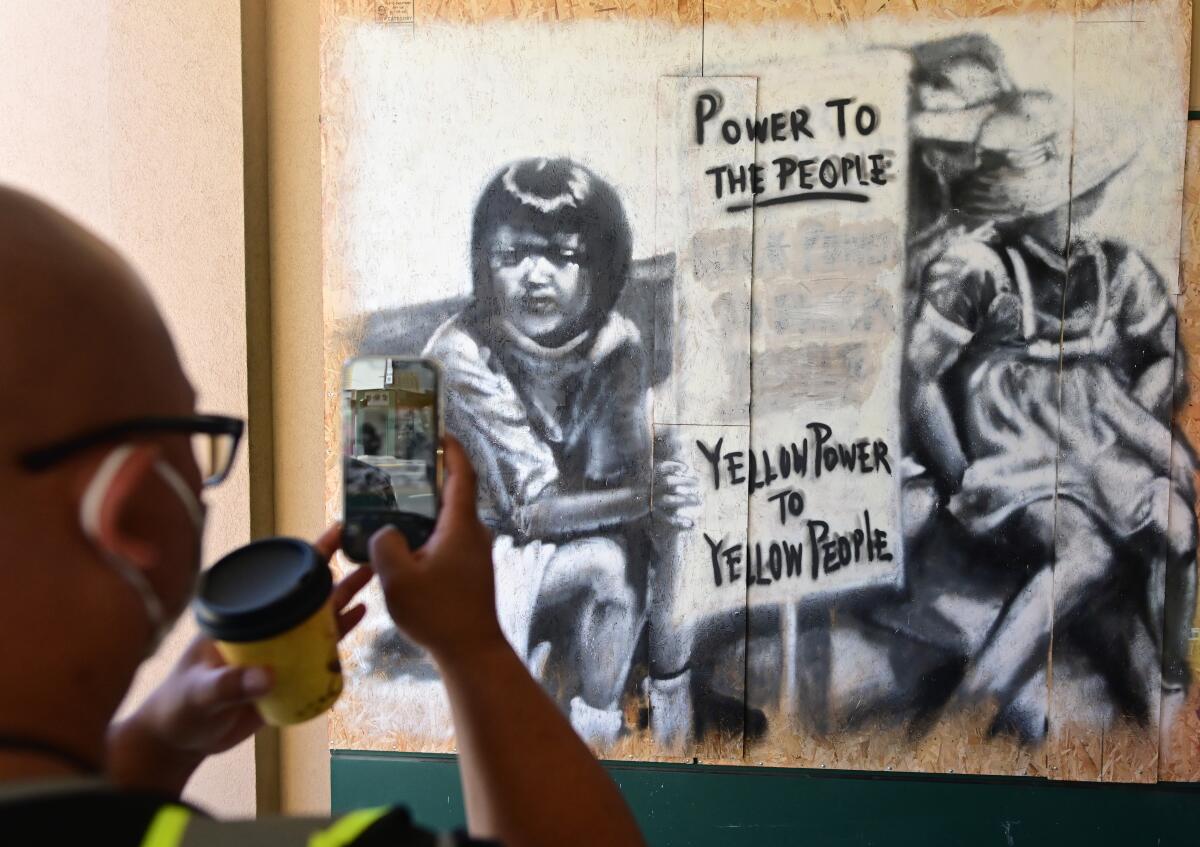 A man uses his phone to take a picture of a mural with the words Power to the people, yellow power to yellow people
