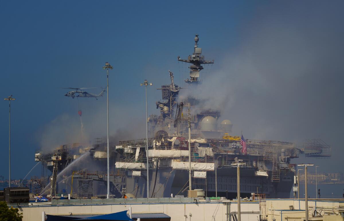 Smoke rises from a large Navy ship as a helicopter drops water and fire tankers spray water