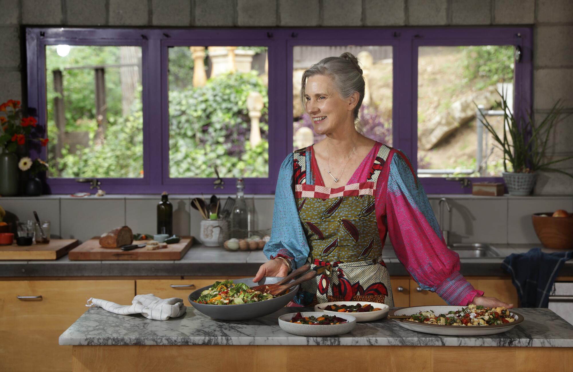 A woman stands in a kitchen, windows behind and salads in big bowls on the counter in front of her
