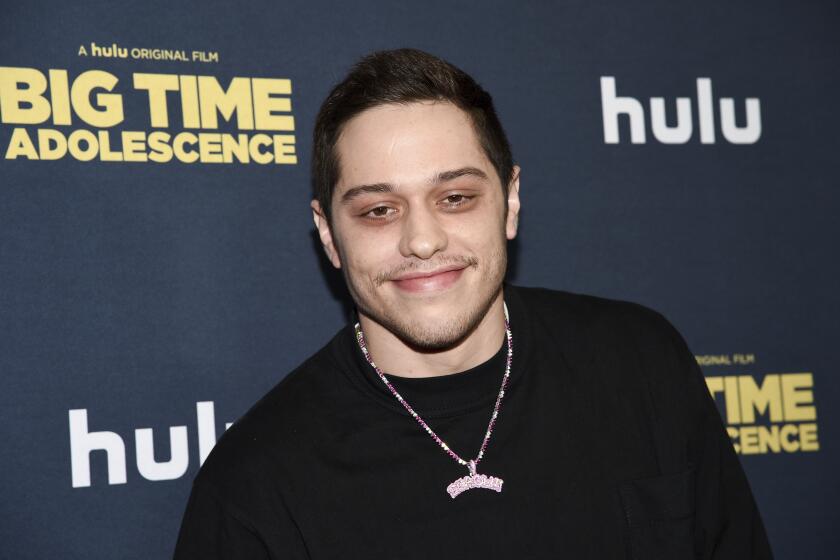 Pete Davidson is smiling and posing while wearing a dark shirt and pink necklace