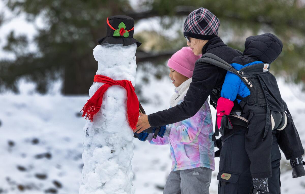 A young girl and her mother build a thin snowman with a hat and scarf.