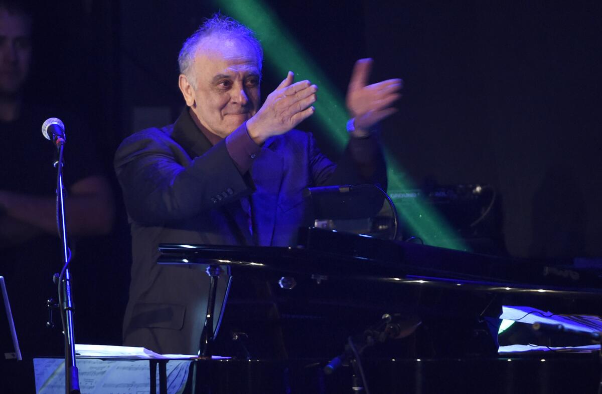Angelo Badalamenti, in a dark blazer and brown shirt gestures, with his hands as he stands on a conductor's podium