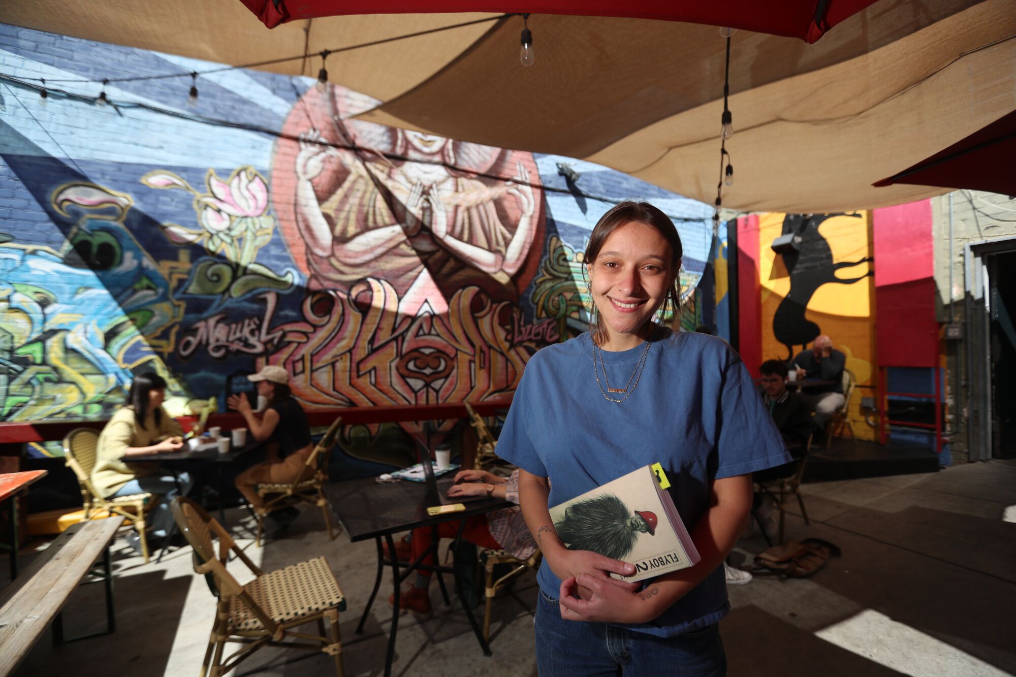 A woman holds a book on an outdoor patio with a mural in the background.