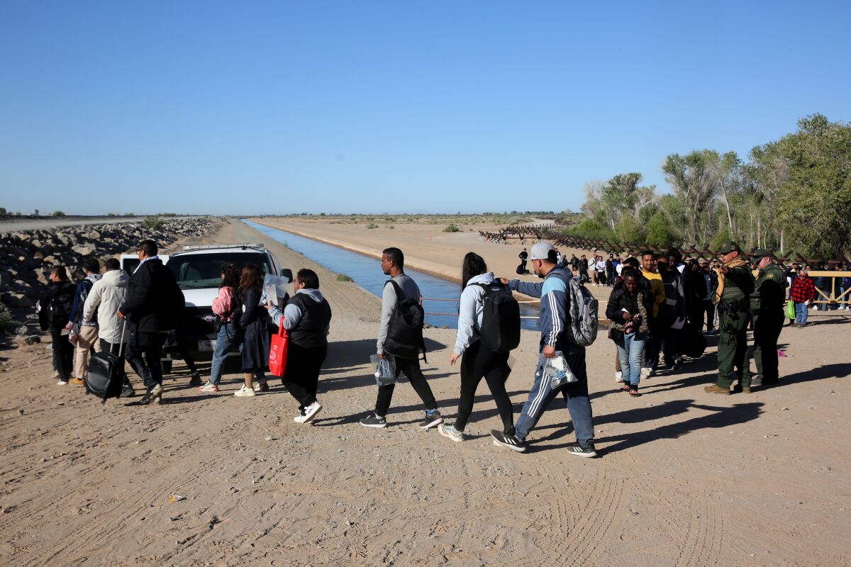 People walking in the dusty earth toward a vehicle as others wait in a long line between trees and a shallow waterway