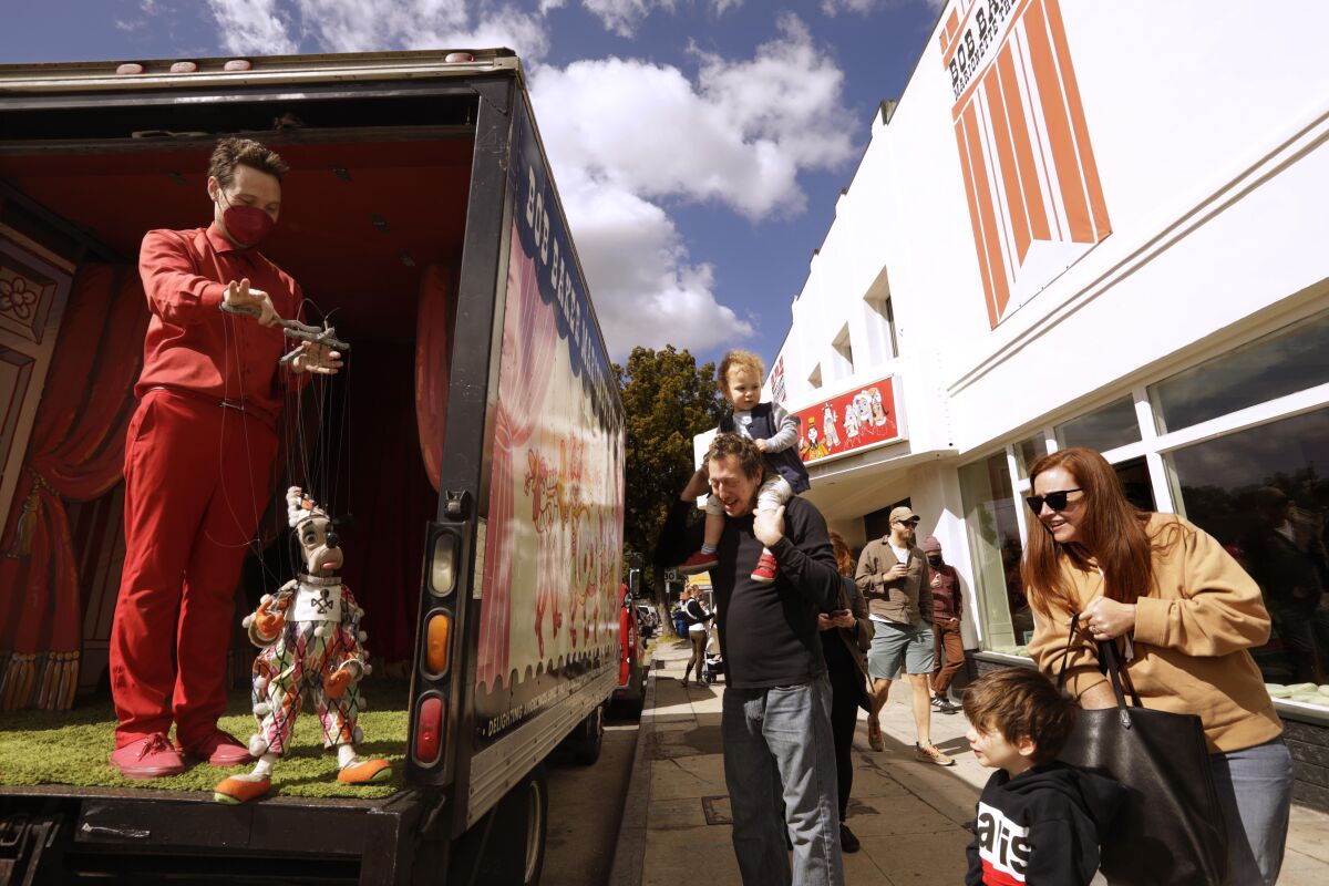 A man stands in the back of a truck operating a marionette. A man with a child on his shoulders stands nearby.