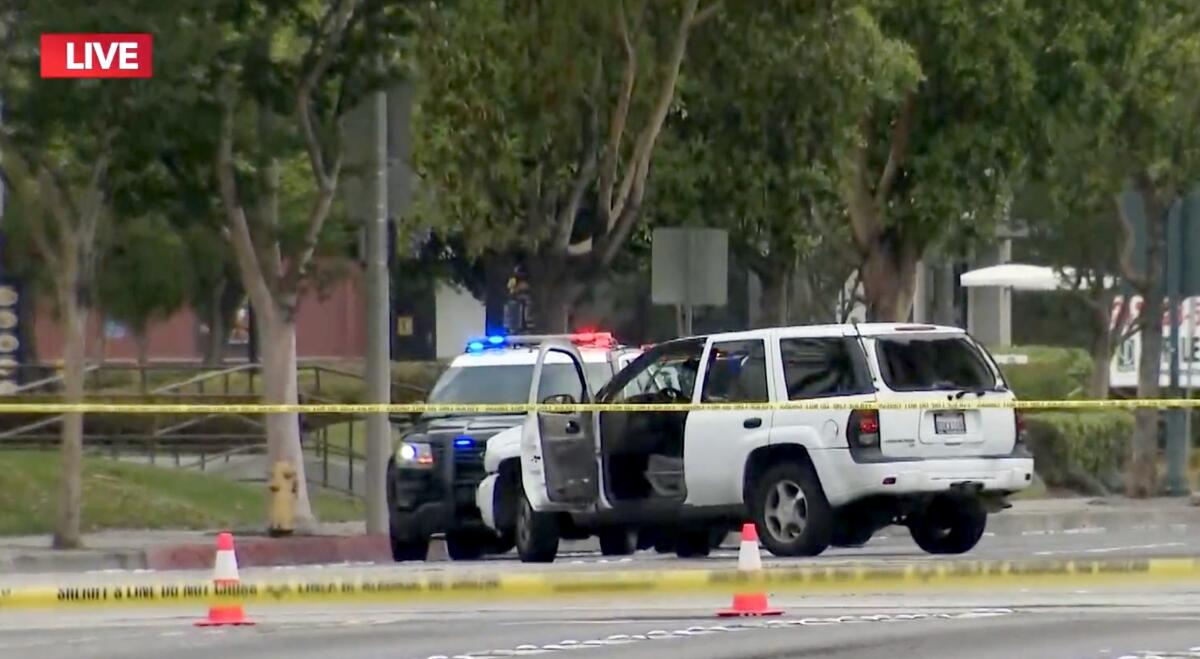 An SUV with its door open and windows blown out in a street behind police tape with a patrol cruiser nearby