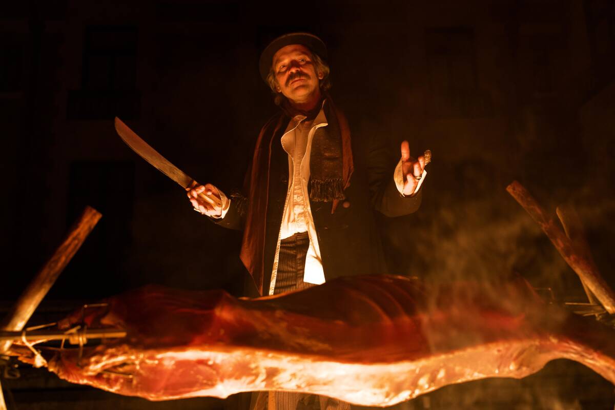 A cook with a knife smiles over a carcass on a spit.