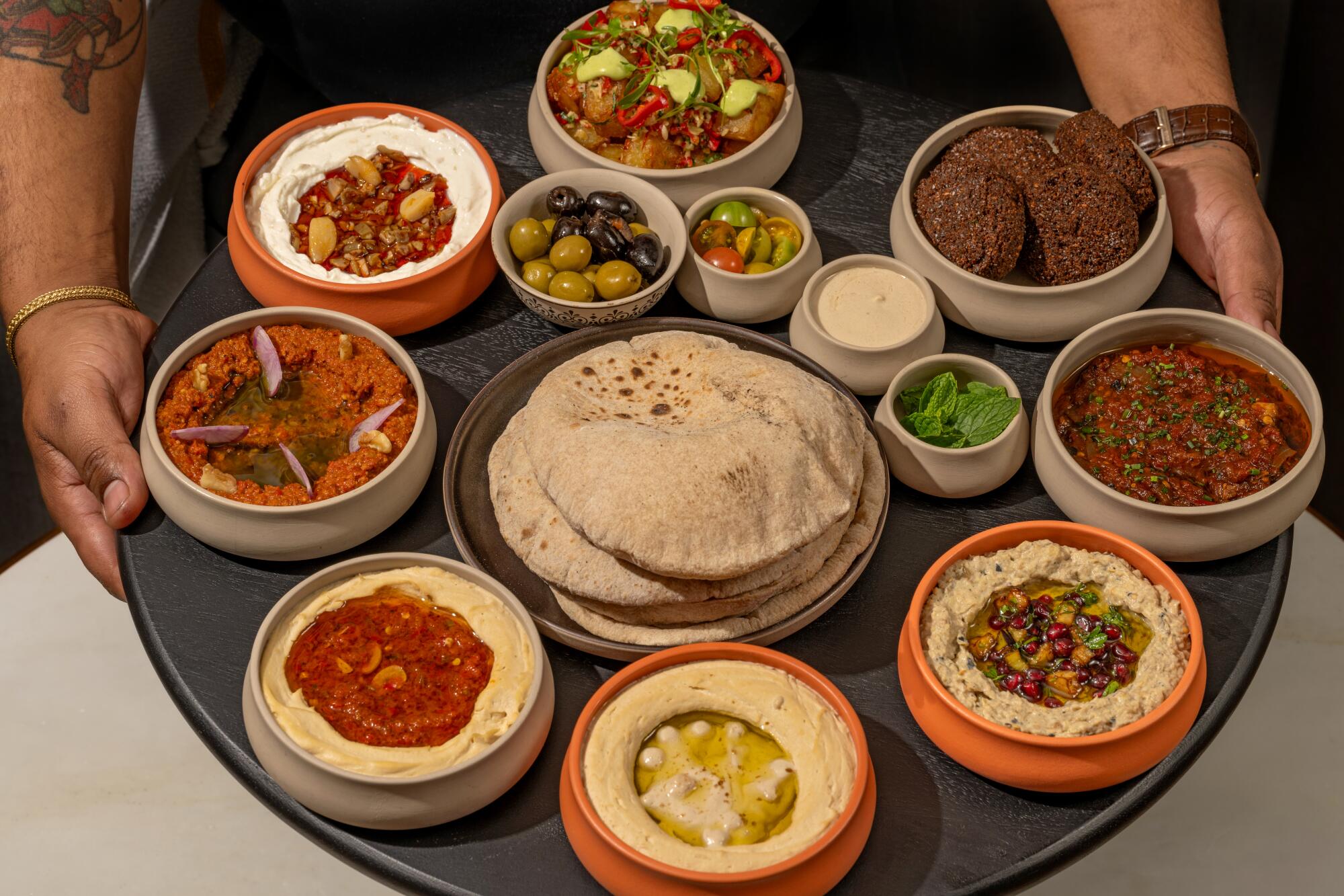 A person holding a large, round platter of round dishes containing an array of Lebanese food.