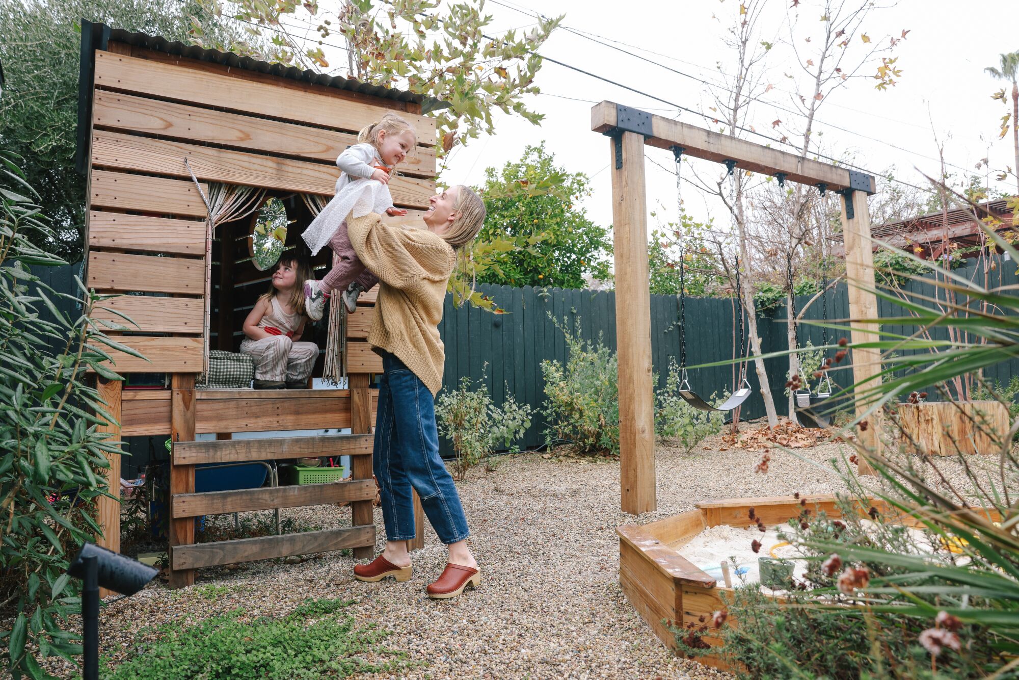 A mother lifts her daughter into an outdoor wooden playhouse.