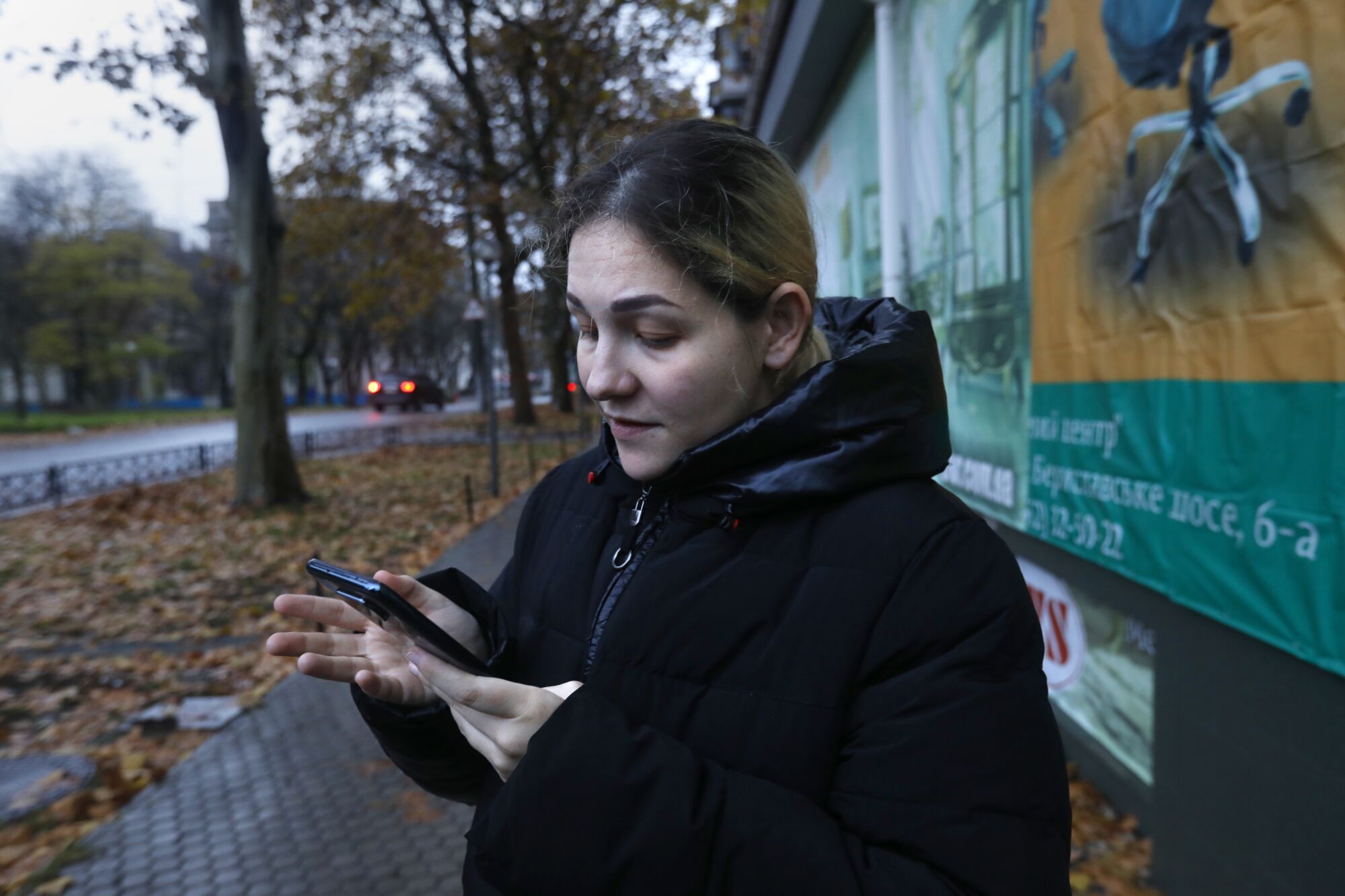 A dark haired woman wearing a black hooded jacket looks at her phone along a tree lined street 