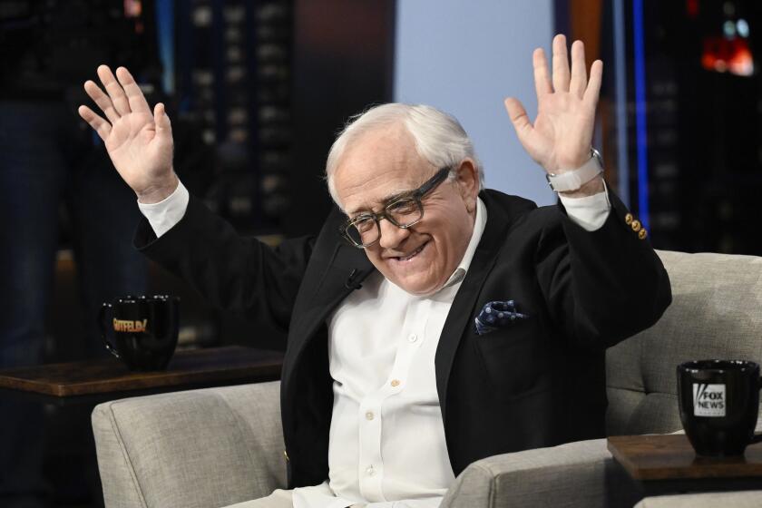 A man with white hair wearing a suit, sitting in an armchair and raising his hands into the air