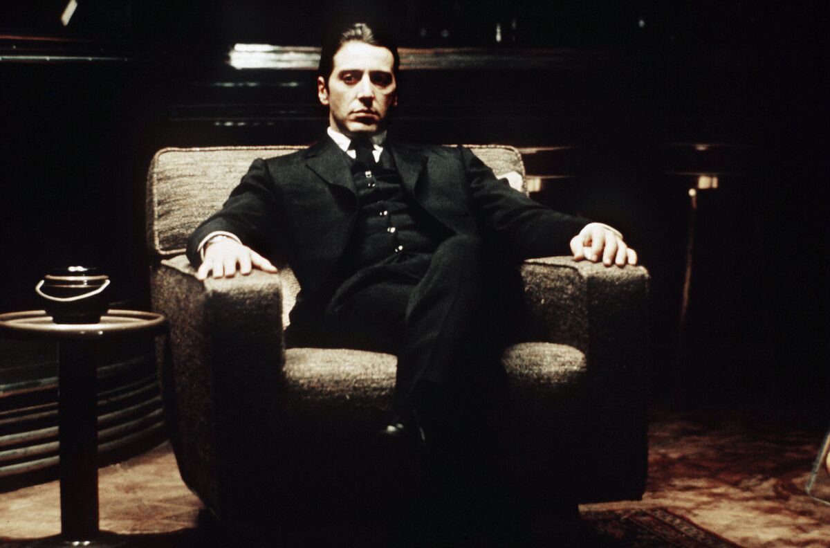 A man with slicked-back dark hair wears a dark three piece suit and sits in an easy chair in a dimly lit room.