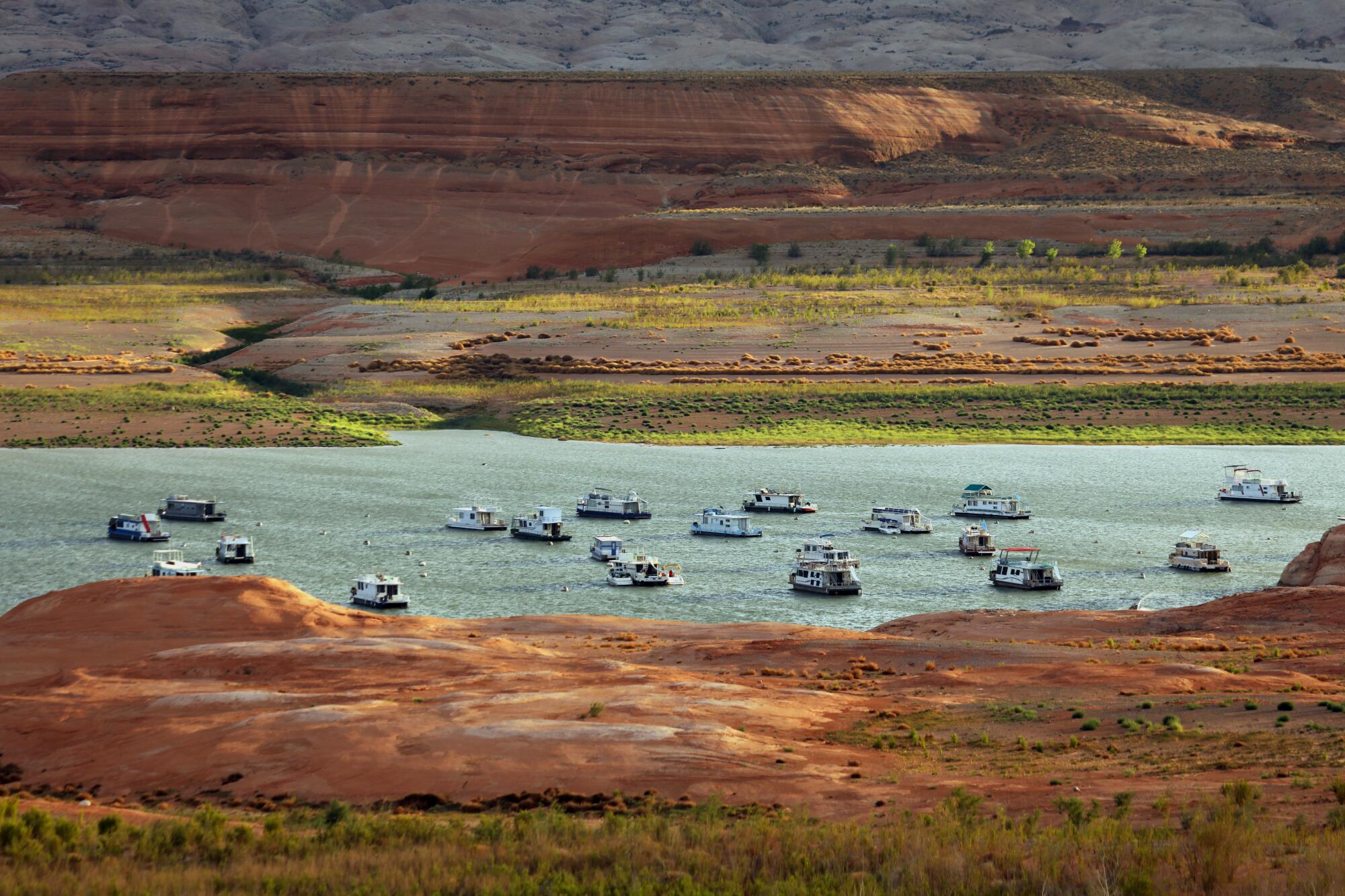 Houseboats seen from a distance dotting a blue-gray lake surrounded by red terrain streaked with green growth
