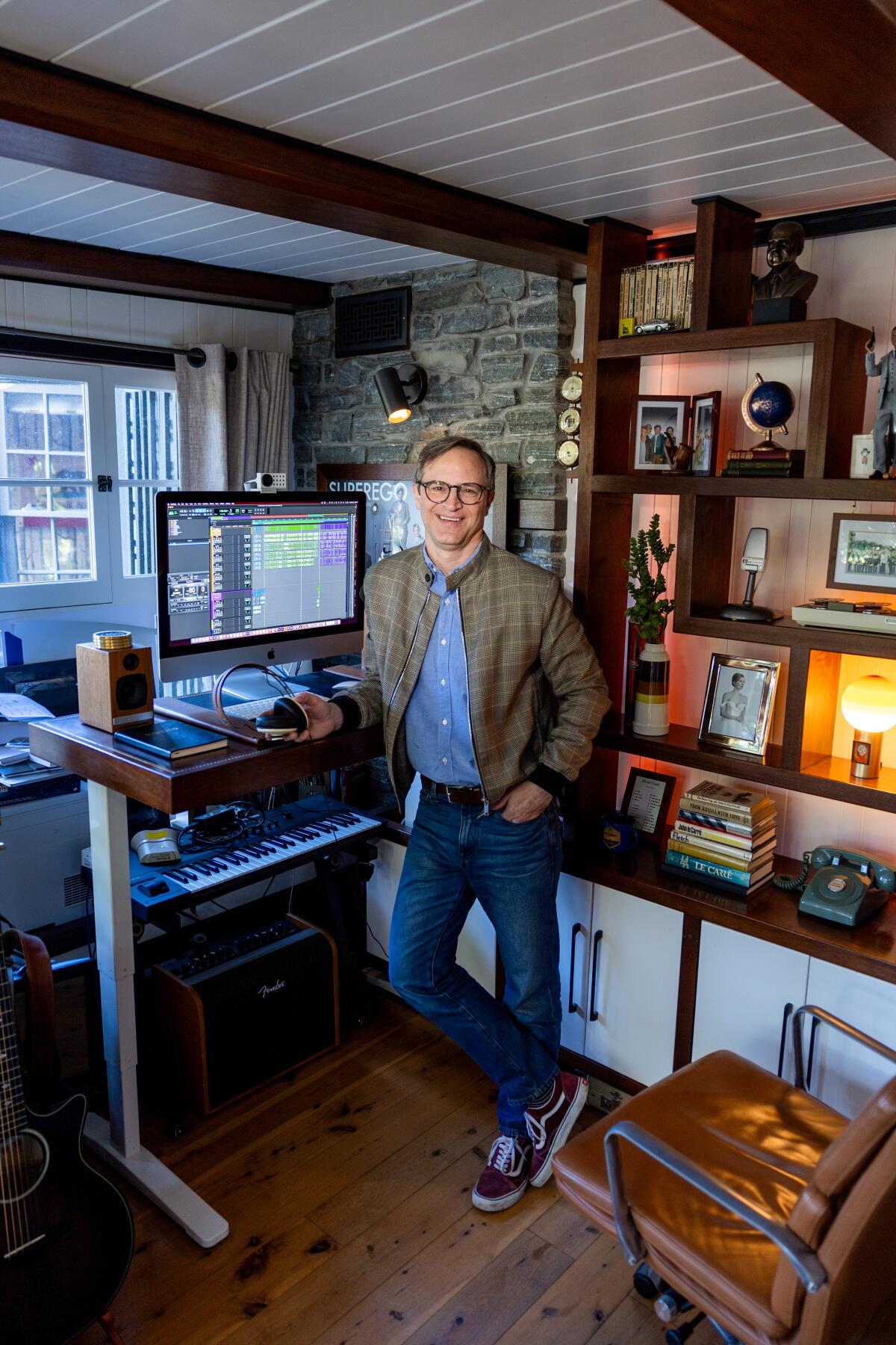 Matt Gourley leans on his standing desk with oversize screen, microphone and wall lined with photos.