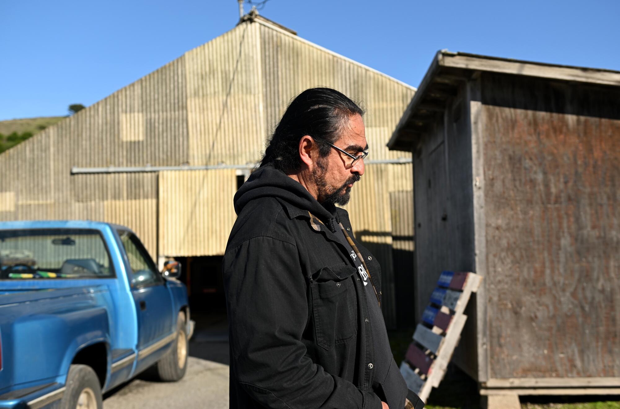 A man stands near a blue pickup truck, a wooden shack and a larger building with a metal roof